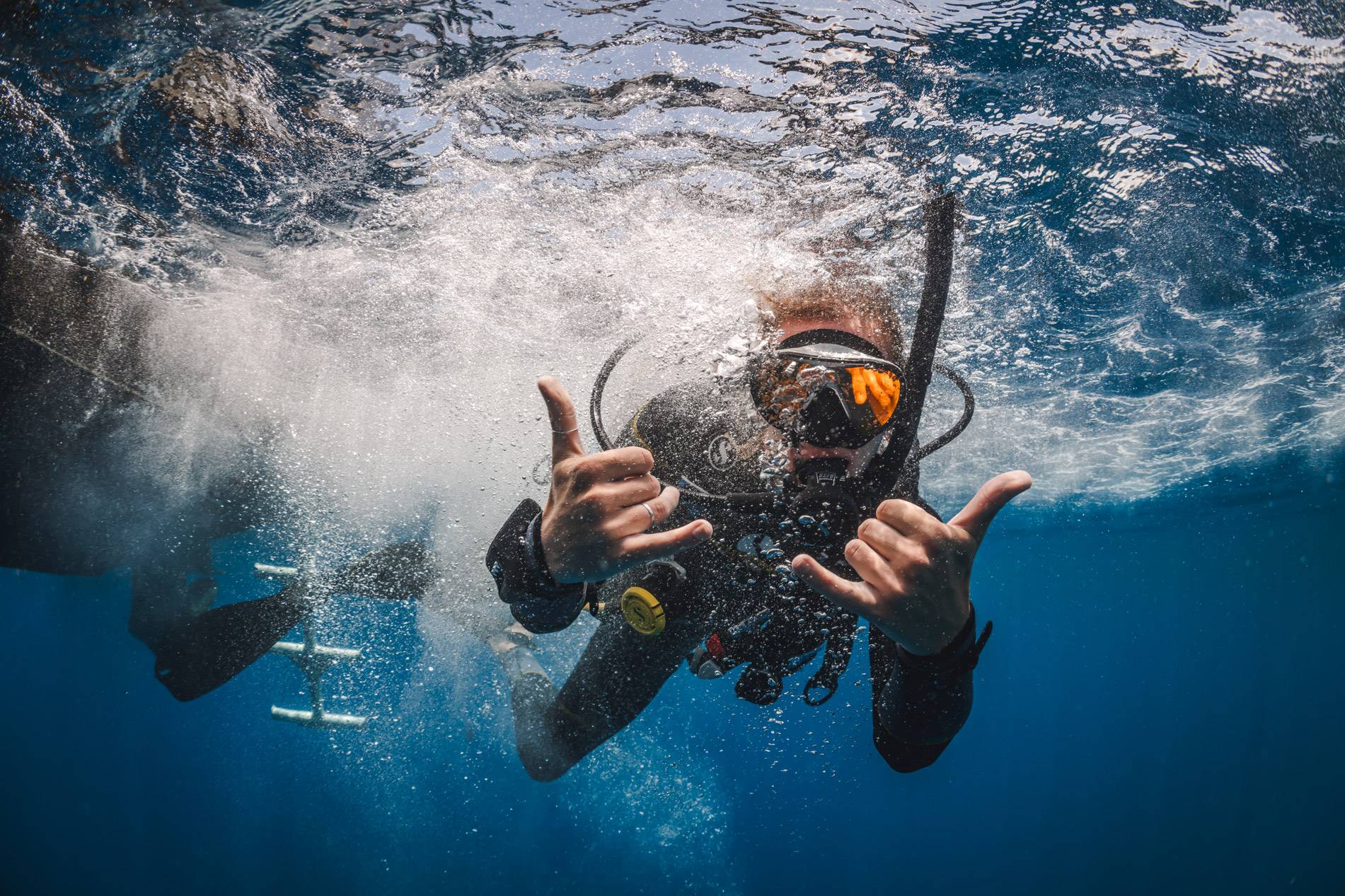 A scuba diver giving a double shaka sign underwater, surrounded by bubbles next to the dive boat